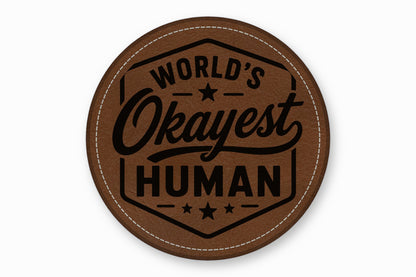 Round brown leatherette coaster featuring the black text World's Okayest Human inside a hexagonal border.