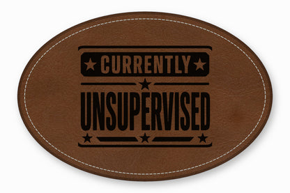 An oval brown leather patch with white stitched borders and black debossed text reading CURRENTLY UNSUPERVISED.
