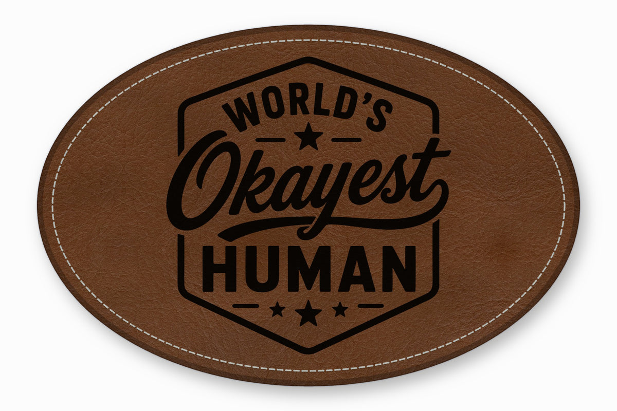 Brown oval leather patch with white stitched border and the engraved text Worlds Okayest Human.