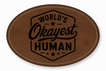 Brown oval leather patch with white stitched border and the engraved text Worlds Okayest Human.