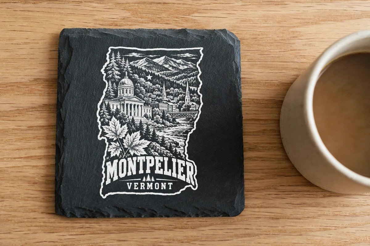 A square black slate coaster engraved with a Montpelier, Vermont cityscape and map outline sits on a wooden surface beside a coffee mug.