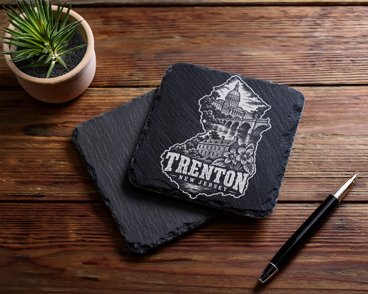 Two black square slate coasters with a laser-engraved Trenton, New Jersey design rest on a wooden table.