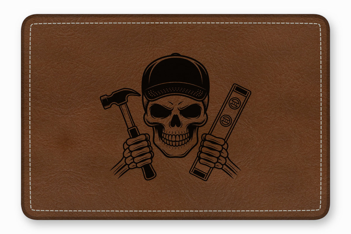 Rectangular brown leather patch with white stitching featuring an engraved skull wearing a cap, holding a hammer and level.