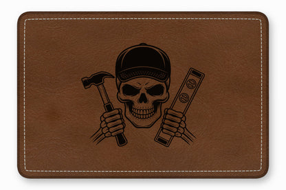 Rectangular brown leather patch with white stitching featuring an engraved skull wearing a cap, holding a hammer and level.