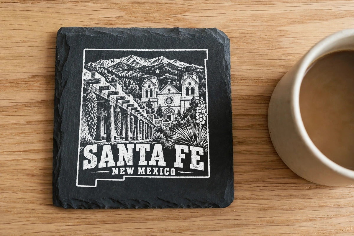 A square slate coaster with a white laser-engraved Santa Fe, New Mexico landscape design rests on a wooden table beside a coffee mug.