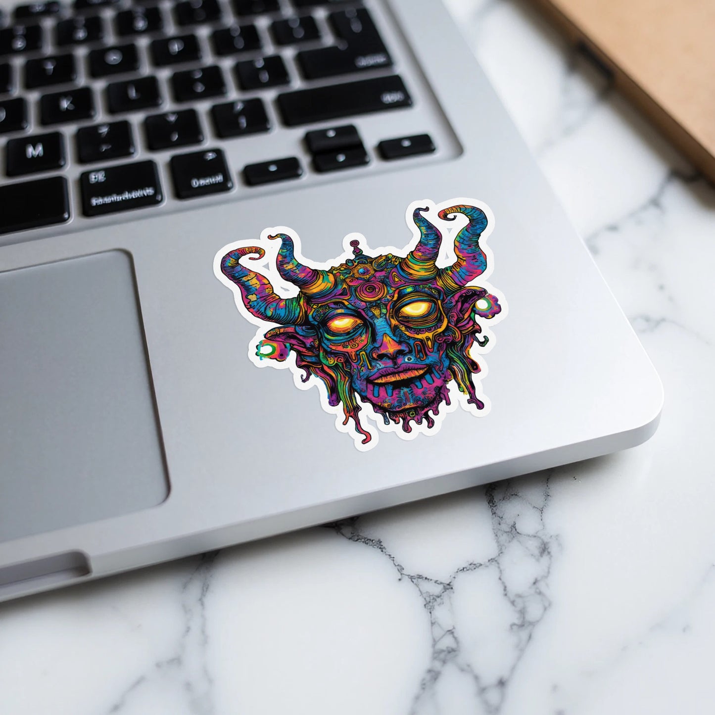 Psychedelic demon face sticker with horns and glowing yellow eyes on a laptop.
