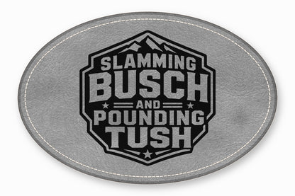 An oval grey leather patch with white stitching featuring a black emblem with the text Slamming Busch and Pounding Tush.