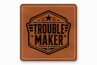 Square brown leather patch with white stitched border and a black emblem reading TROUBLE MAKER.