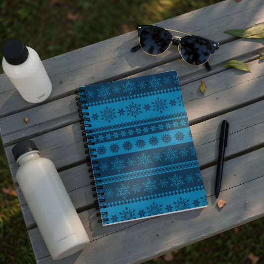 Spiral notebook with blue snowflake pattern on wooden bench with water bottles and sunglasses.
