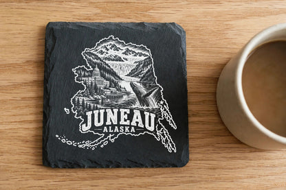 A square slate coaster engraved with a Juneau Alaska map, mountains, and whale sits on a wooden table.