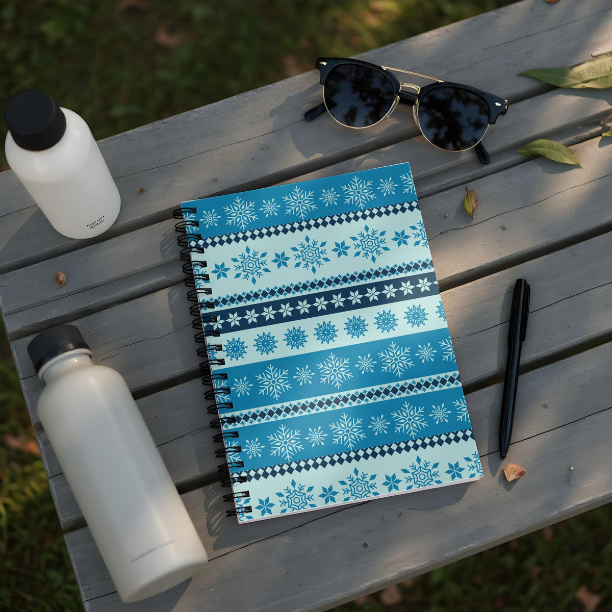 Notebook with snowflake pattern on a wooden bench with sunglasses and water bottles.