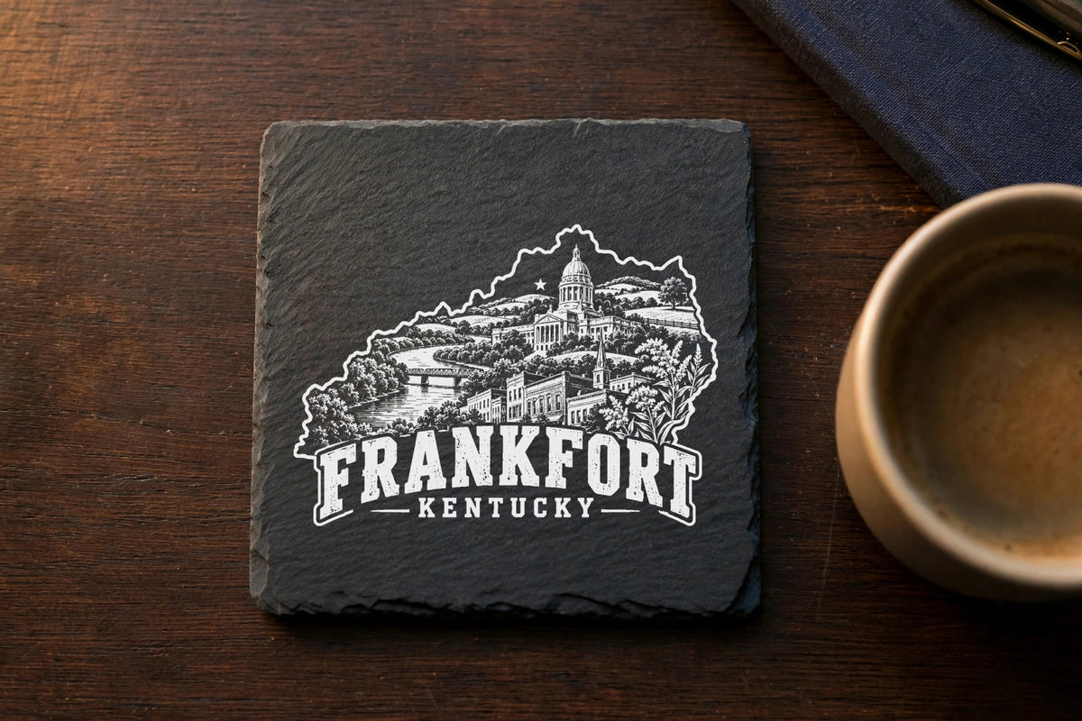 Square black slate coaster engraved with a white illustration of Frankfort, Kentucky, on a wooden table.