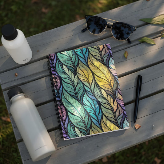 Notebook with feather pattern, two water bottles, sunglasses, and pen on wooden bench.