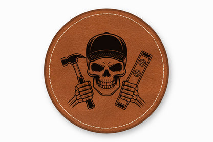 A round brown leather patch with white stitching features a skull wearing a cap, holding a hammer and level.