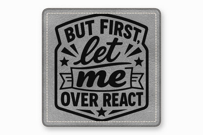 A square gray leather patch featuring black text that reads, "BUT FIRST, let me OVER REACT".