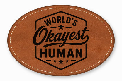 An oval brown leather patch with white stitched edges and black text reading World's Okayest Human.