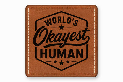 Brown square leather patch with stitched borders and the text Worlds Okayest Human inside a hexagon.