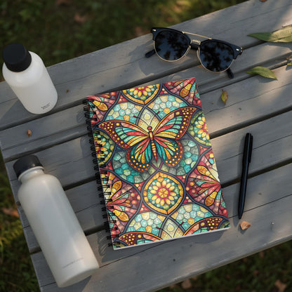 Spiral notebook with stained-glass butterfly design on wooden bench with water bottles.