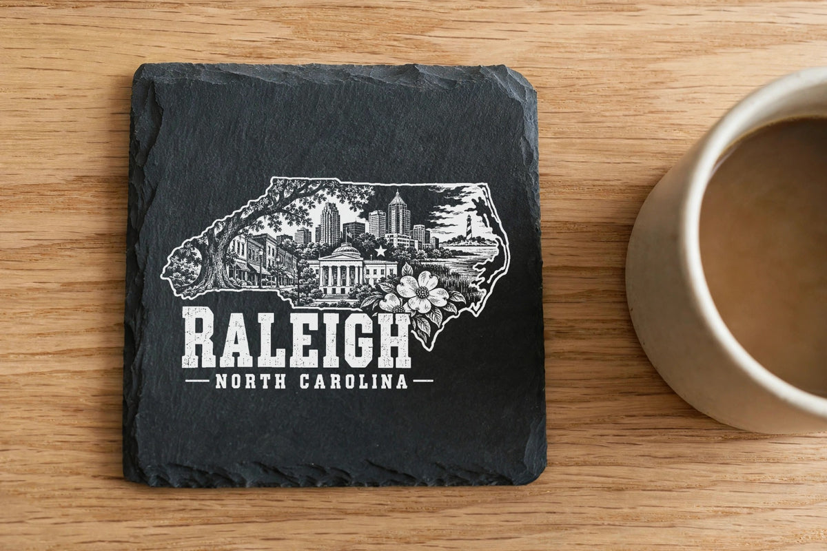Square black slate coaster engraved with a Raleigh, North Carolina skyline design next to a coffee mug.