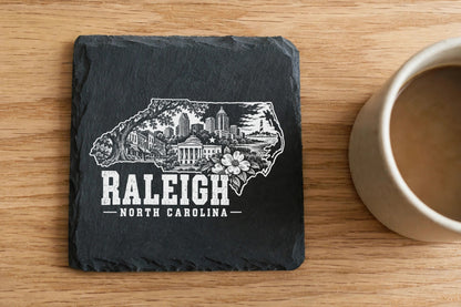 Square black slate coaster engraved with a Raleigh, North Carolina skyline design next to a coffee mug.