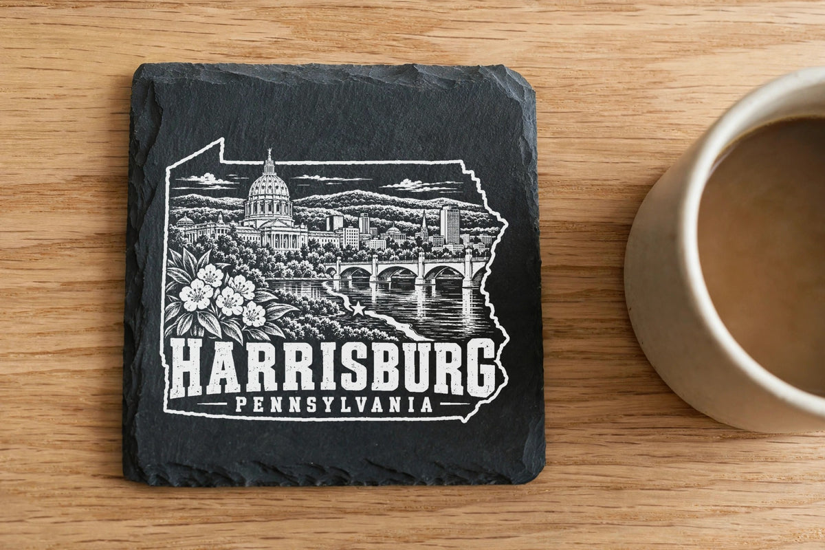 Square black slate coaster engraved with a white illustration of Harrisburg, Pennsylvania, sitting on a wooden surface next to a coffee cup.