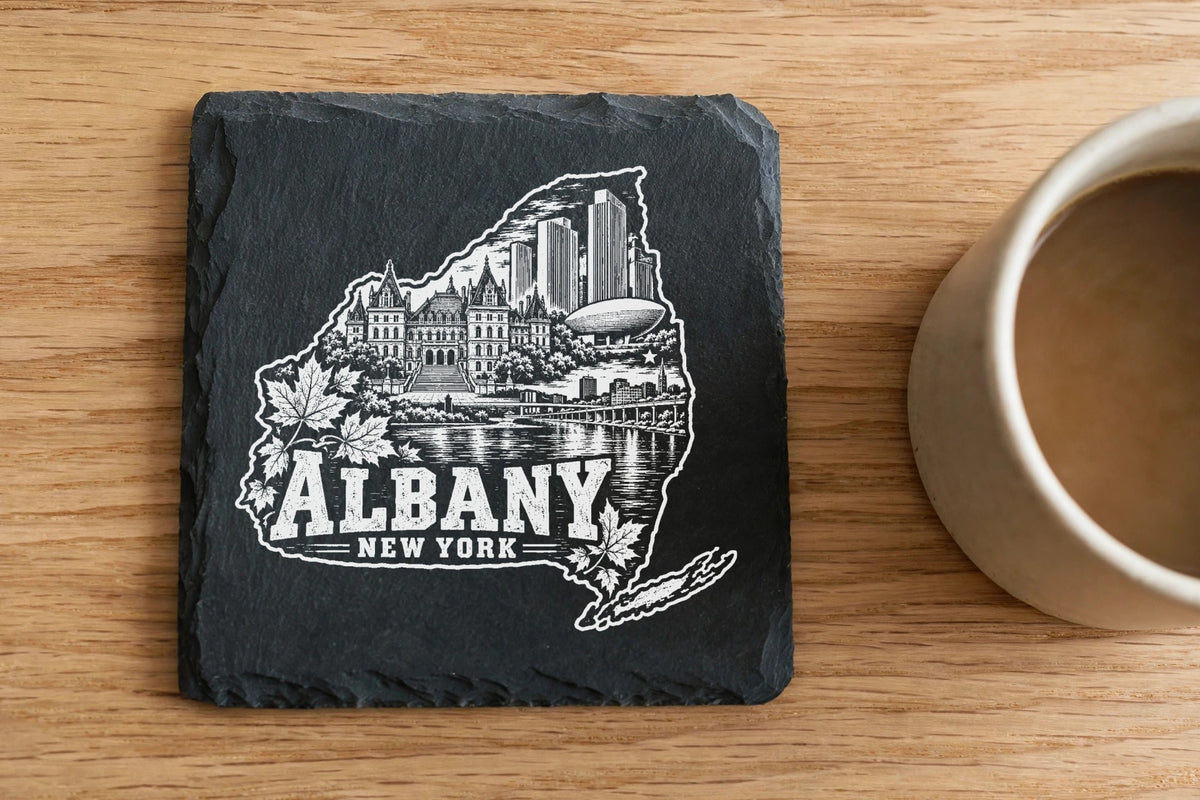 Square black slate coaster engraved with an Albany New York skyline map beside a coffee mug.