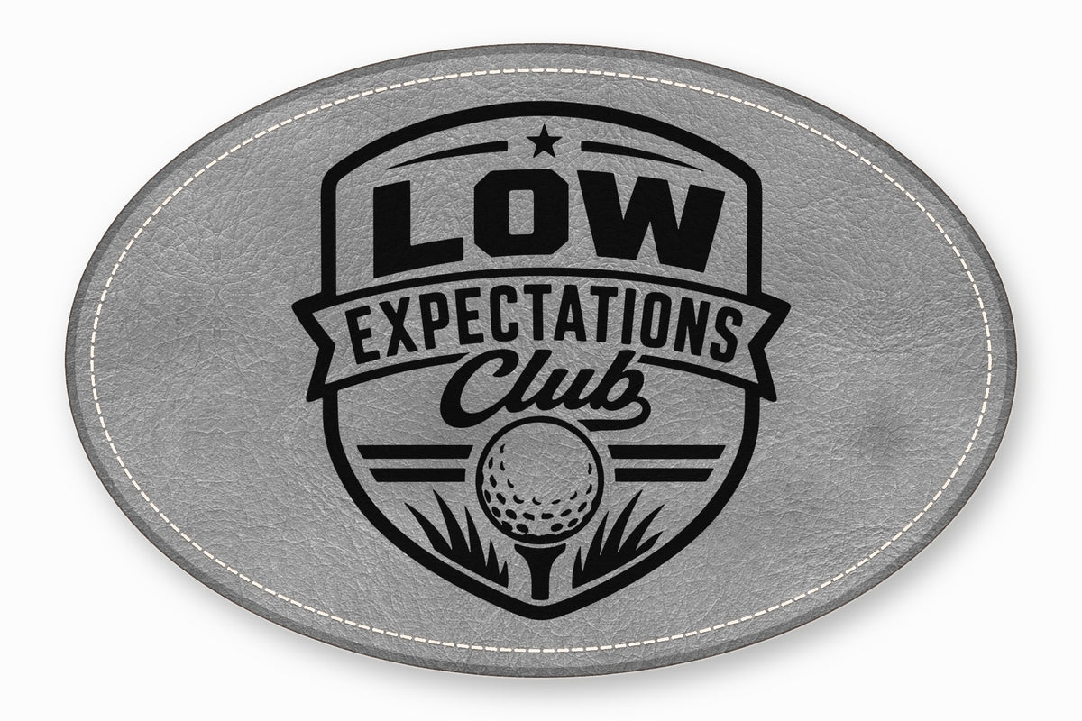 An oval grey leather patch with white stitched edges and a black golf emblem featuring the text Low Expectations Club.