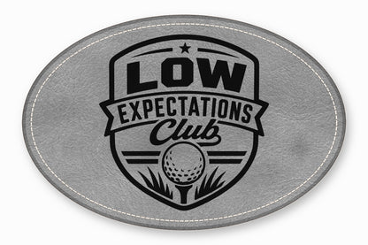 An oval grey leather patch with white stitched edges and a black golf emblem featuring the text Low Expectations Club.