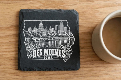 A square black slate coaster engraved with a Des Moines skyline illustration rests on a wooden table beside a coffee mug.