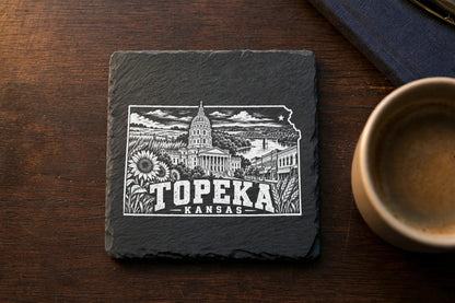 Square slate coaster engraved with a white illustration of Topeka, Kansas landmarks, set on a wooden table.