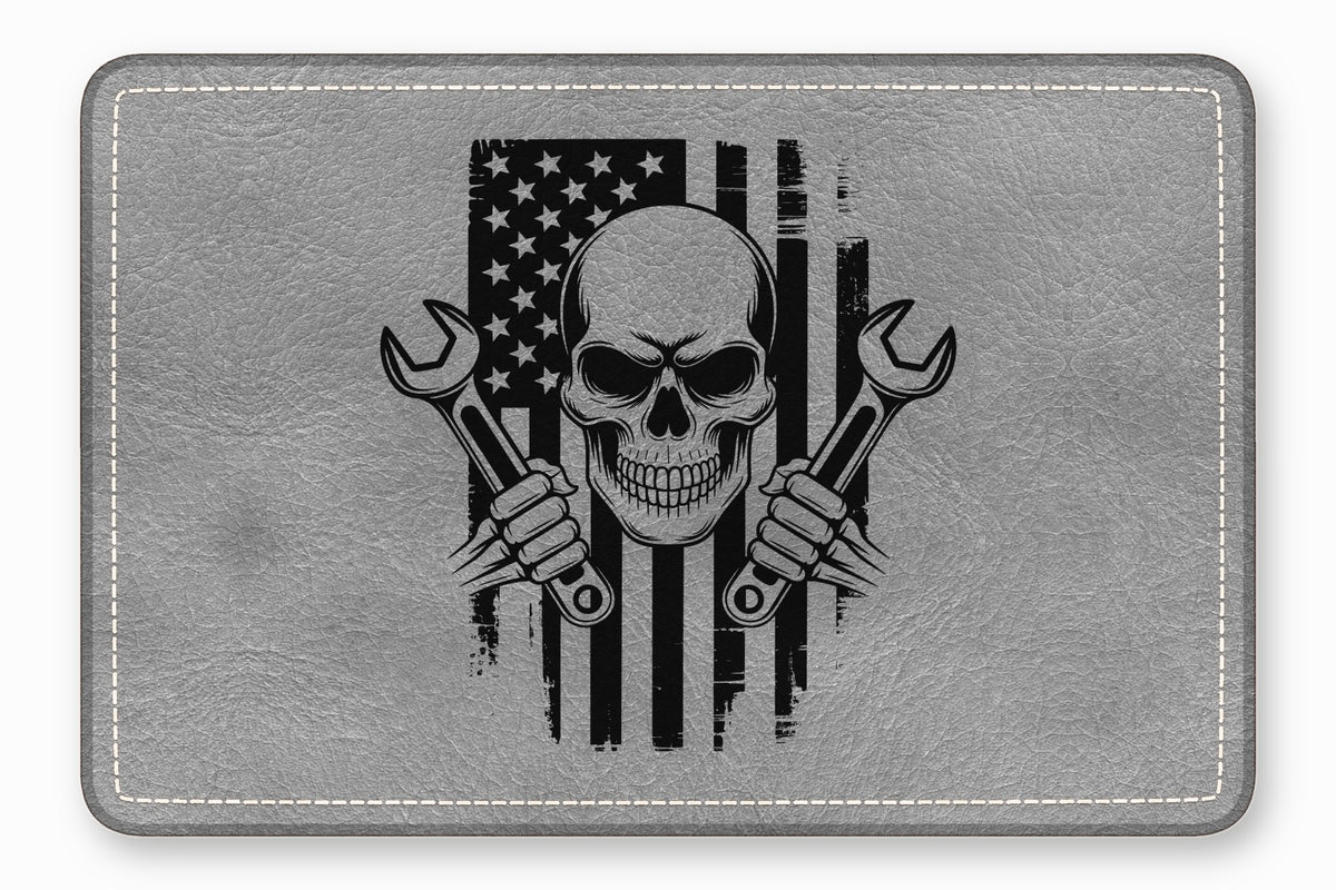 A grey leather patch with rounded corners features a black graphic of a skull holding two crossed wrenches over a distressed American flag.