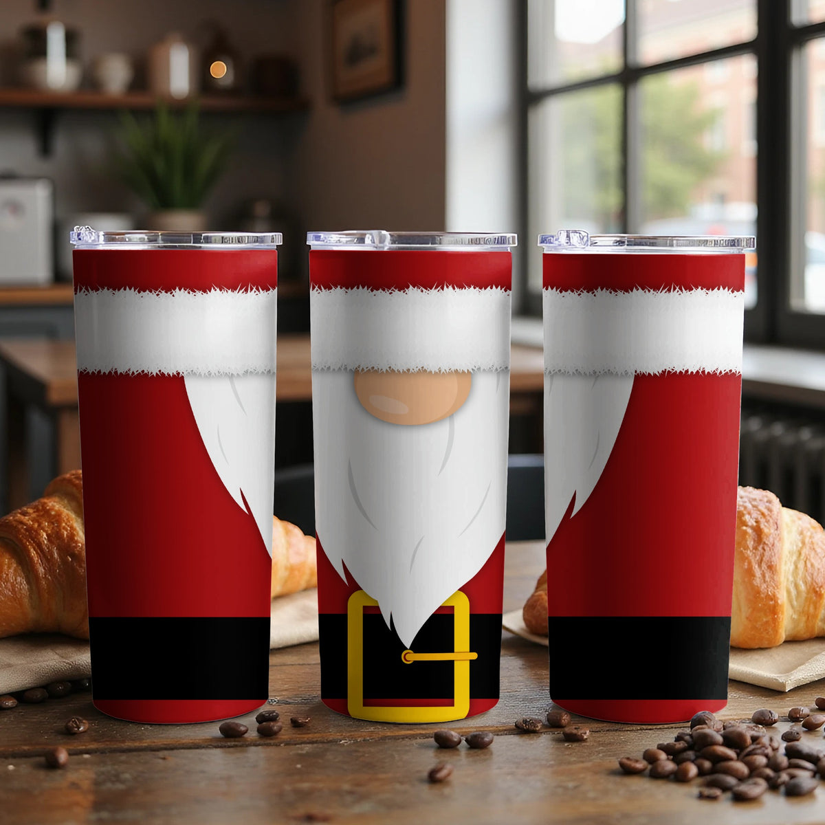 Three Santa-themed tumblers with clear lids sit on a wooden table with croissants and coffee beans.