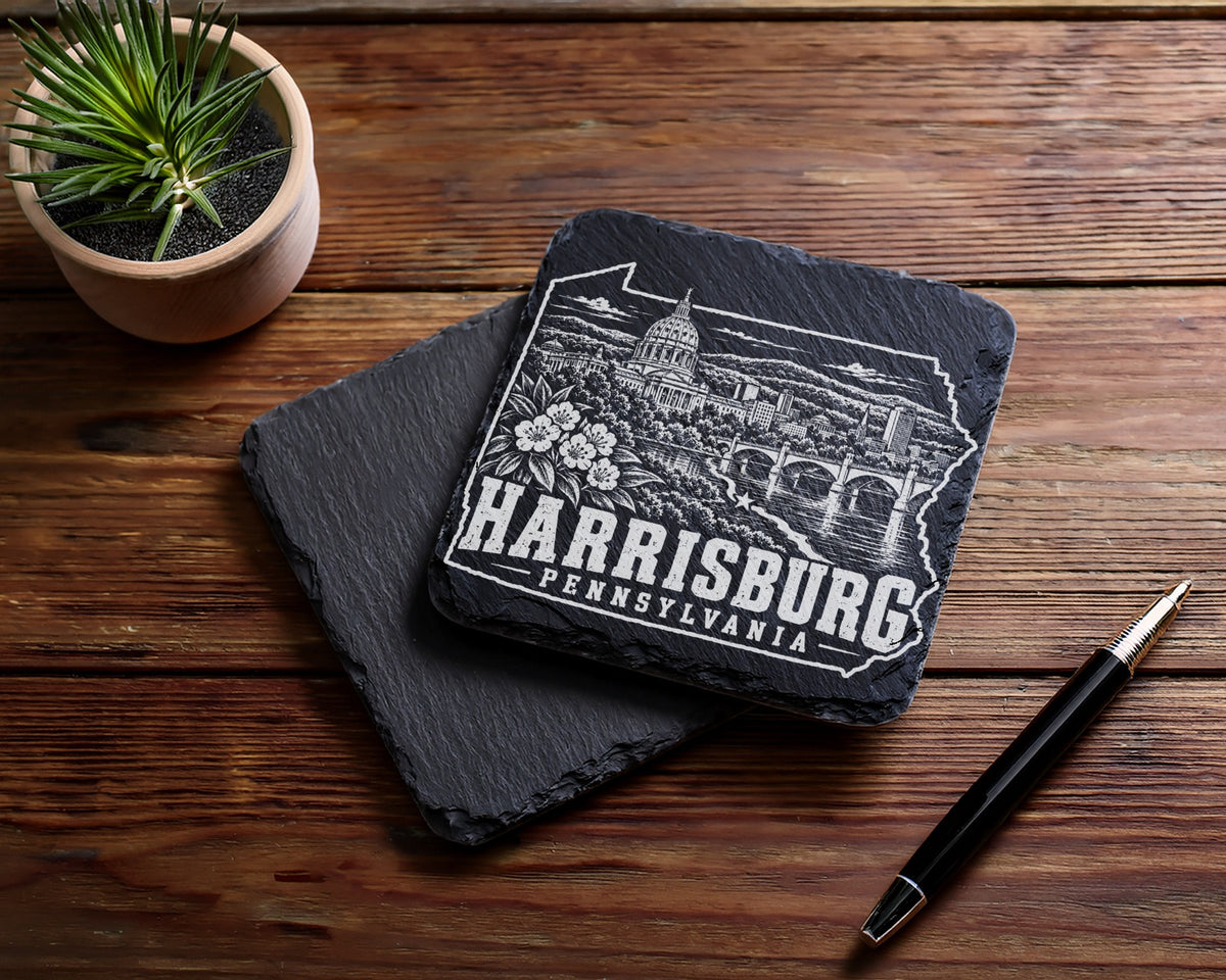 Two square black slate coasters on a wooden surface, engraved with a Harrisburg, Pennsylvania skyline design.