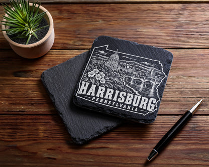 Two square black slate coasters on a wooden surface, engraved with a Harrisburg, Pennsylvania skyline design.