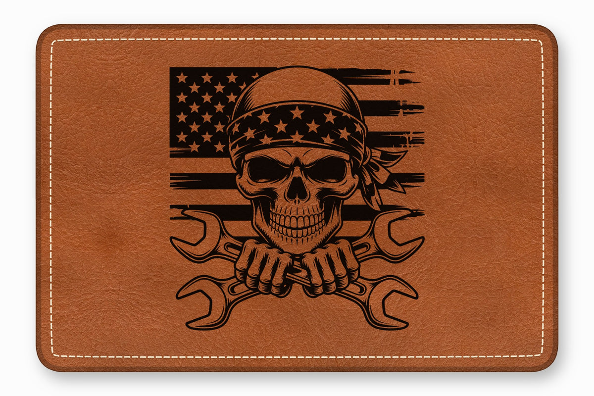 A brown leather patch with white stitched edges features a black engraved skull, American flag, and crossed wrenches.