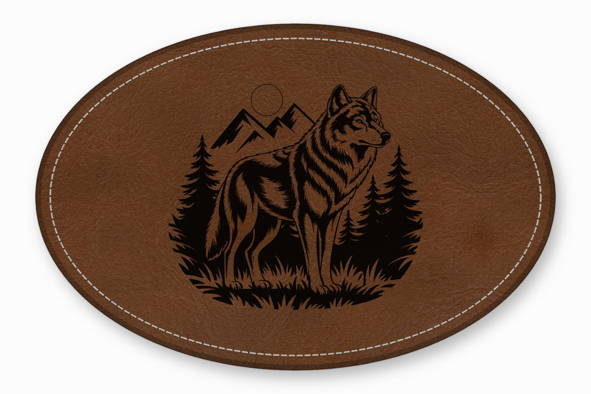 Brown oval leather patch featuring a black engraved wilderness scene with a wolf, trees, and mountains.