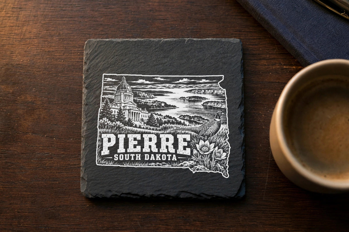 A square slate coaster with a white engraved map of South Dakota featuring Pierre, a pheasant, and local landmarks.