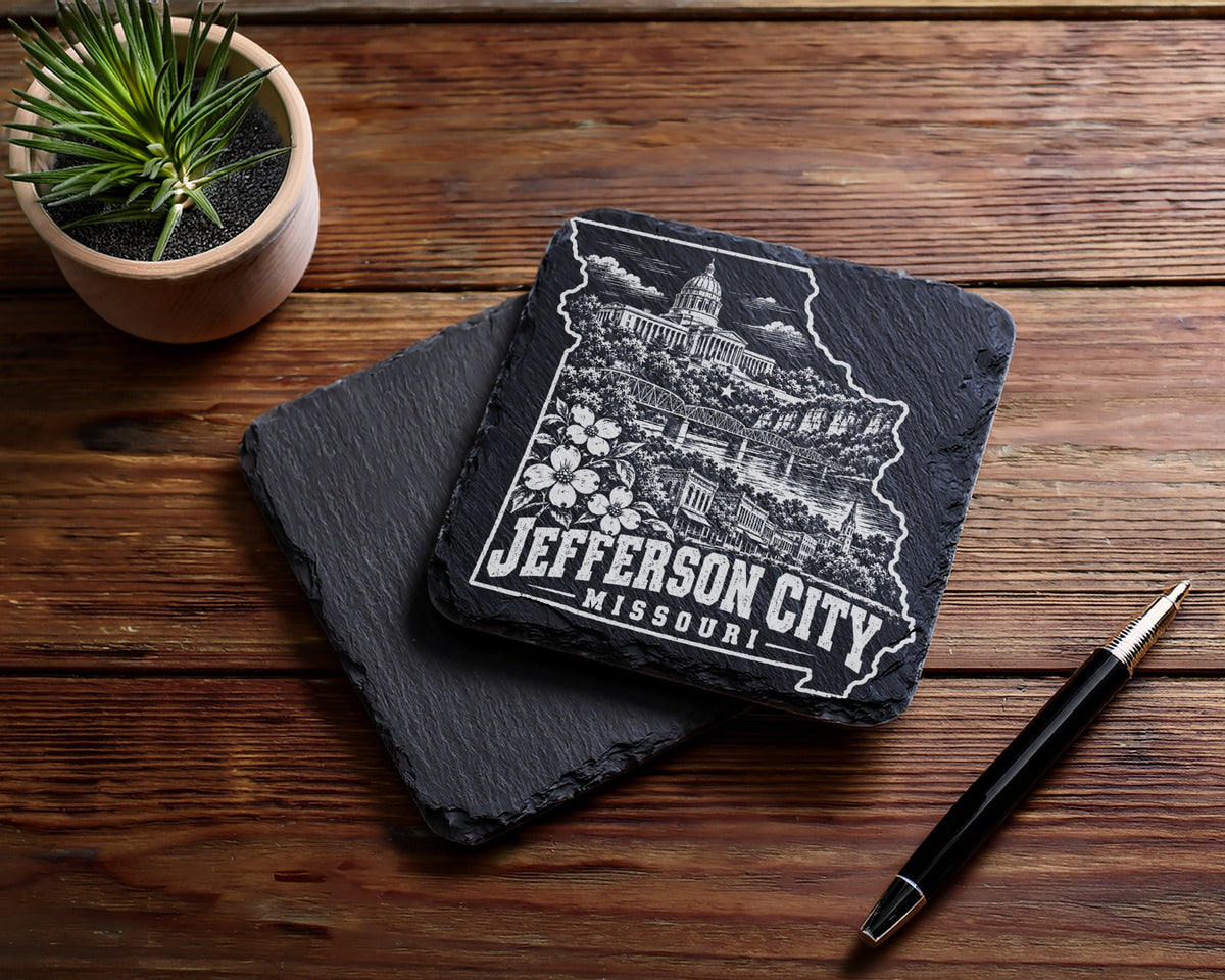 Two square slate coasters rest on a wooden table beside a small potted plant and pen.