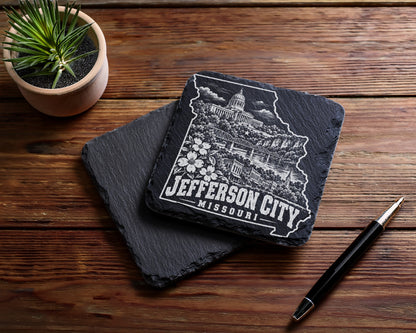 Two square slate coasters rest on a wooden table beside a small potted plant and pen.