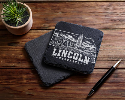Two square slate coasters featuring an engraved Lincoln, Nebraska skyline design sit on a wooden table beside a small potted plant and a pen.