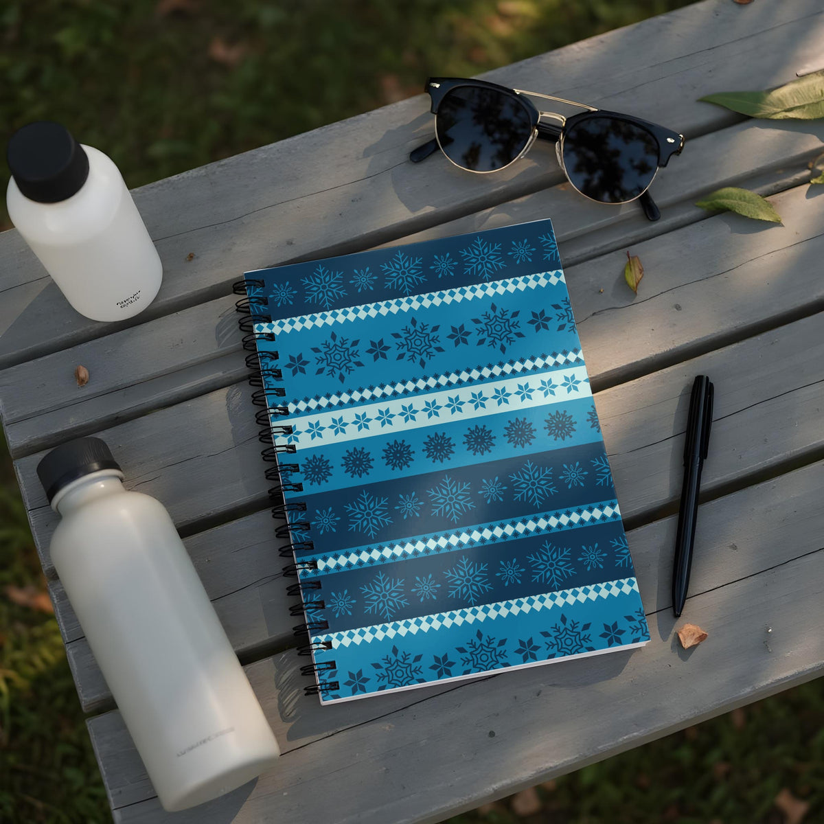 Spiral notebook with blue snowflake pattern on a wooden table with water bottles and sunglasses.