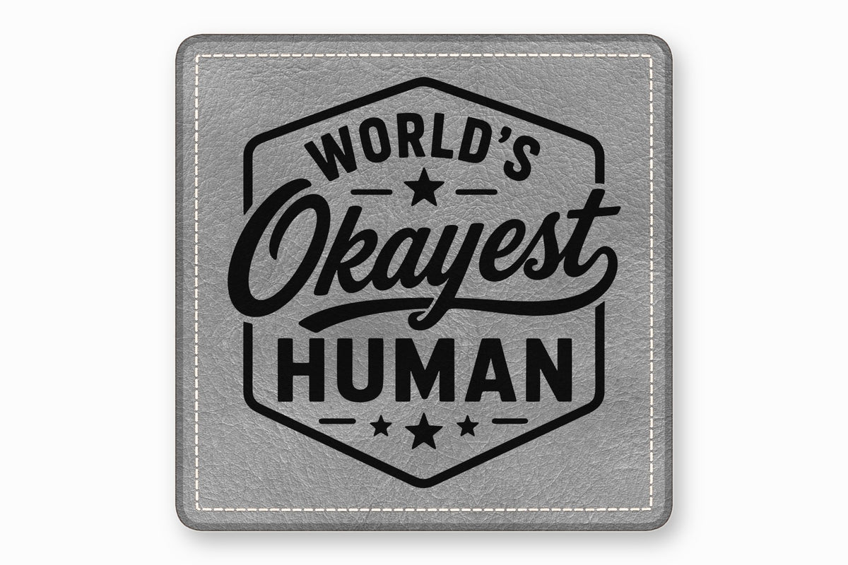 A square grey leather patch with white stitched borders and black text reading Worlds Okayest Human.