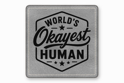 A square grey leather patch with white stitched borders and black text reading Worlds Okayest Human.