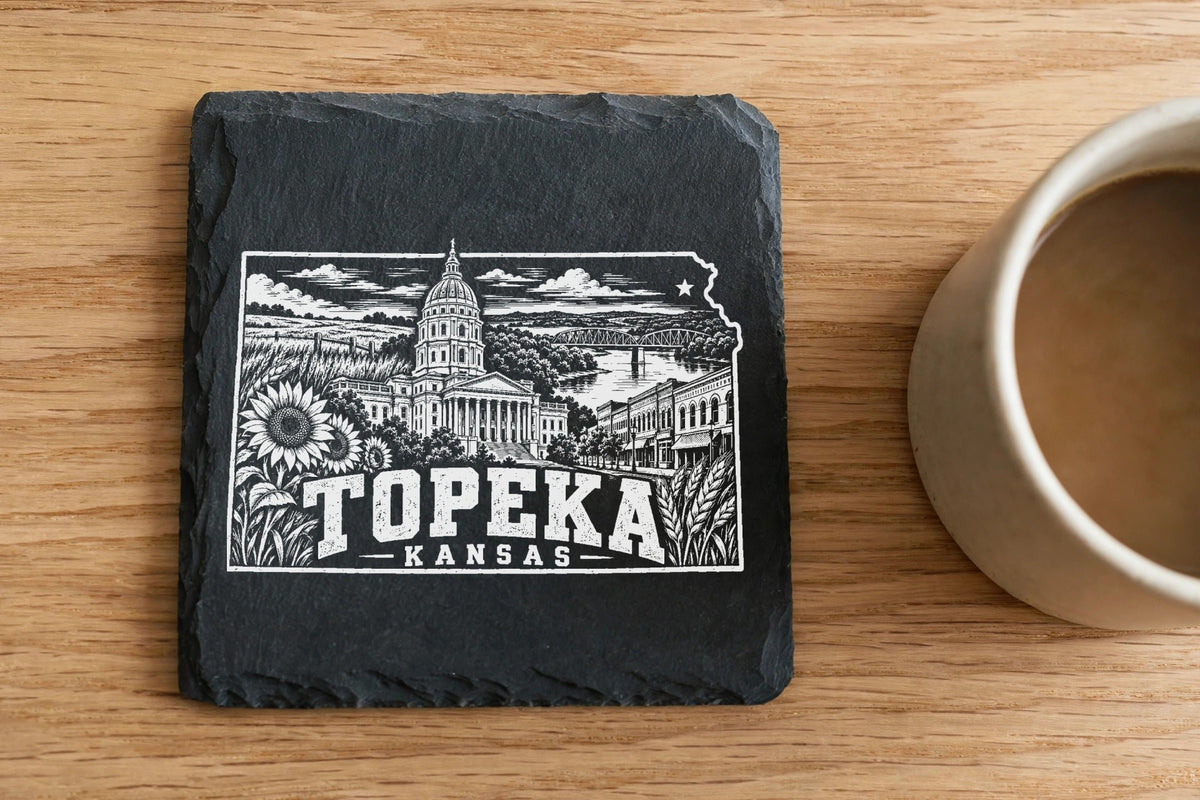 A black square slate coaster with a white Topeka, Kansas engraving sits on a wooden table beside a coffee mug.