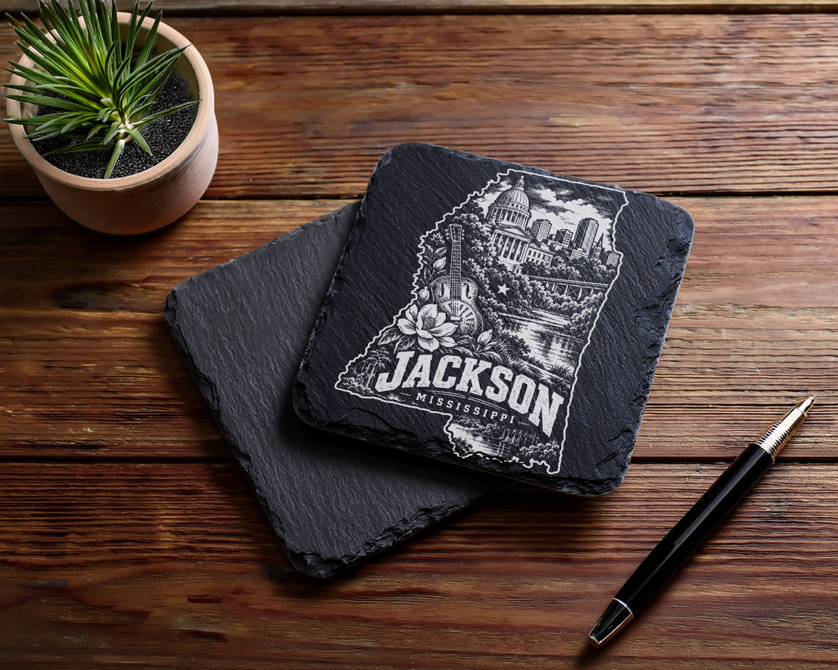 Two black slate coasters resting on a wooden table beside a small potted plant and pen.