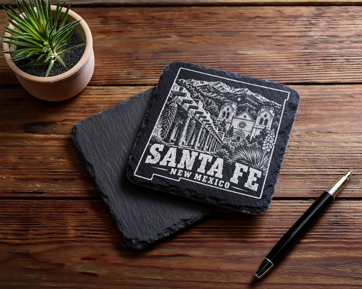 Two square black slate coasters featuring a Santa Fe, New Mexico engraving lie on a wooden table.