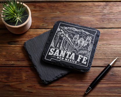 Two square black slate coasters featuring a Santa Fe, New Mexico engraving lie on a wooden table.