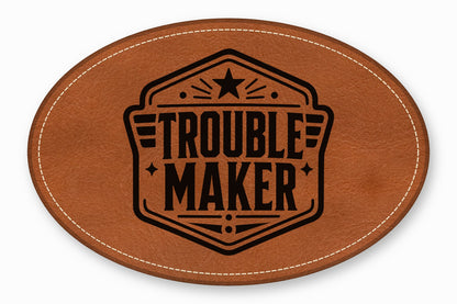 An oval brown leather patch featuring white stitching along the edge and a black printed emblem with the text Trouble Maker.