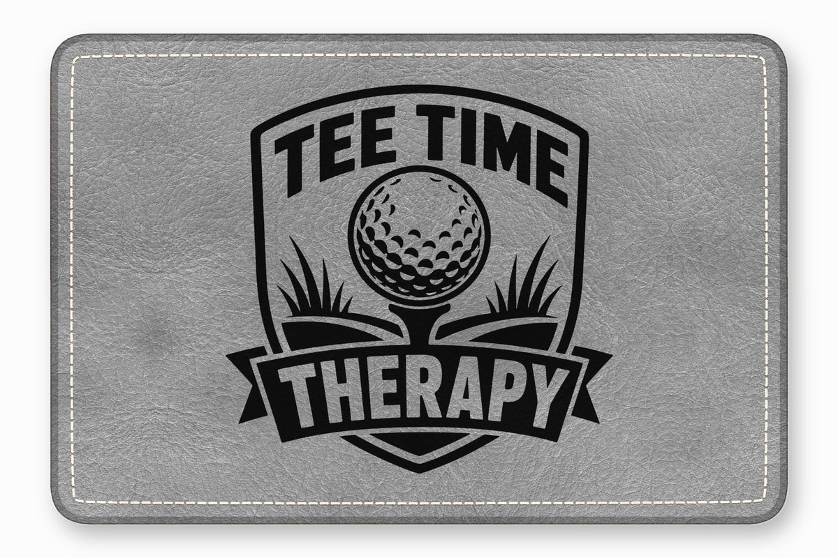 A rectangular gray leatherette patch featuring a black embroidered design with a golf ball and the text Tee Time Therapy.