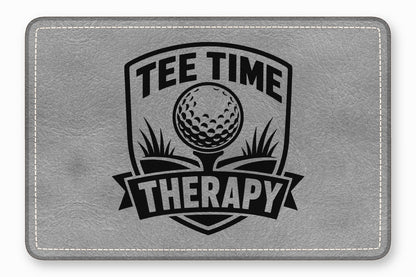 A rectangular gray leatherette patch featuring a black embroidered design with a golf ball and the text Tee Time Therapy.